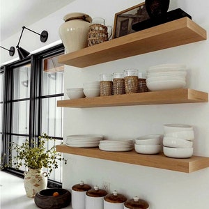 May include: A kitchen interior featuring three wooden shelves against a white wall. The shelves display white bowls, plates, glasses, and decorative jars. A black window frame and a vase with greenery are also visible.