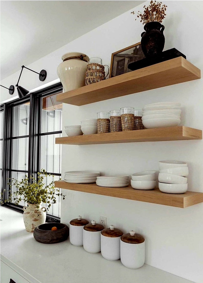 May include: A kitchen interior featuring three wooden shelves against a white wall. The shelves display white bowls, plates, glasses, and decorative jars. A black window frame and a vase with greenery are also visible.