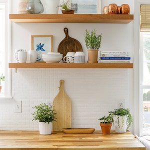 May include: A kitchen scene with wooden shelves displaying various items. Includes plants, framed art, mugs, bowls, cutting boards, and decorative vases. The countertop is wood, and the cabinets are dark blue.