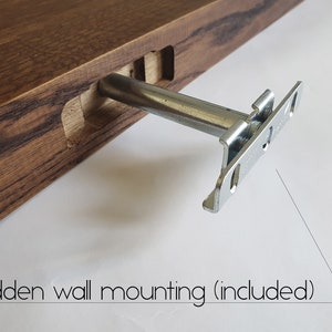 May include: Close-up of a wooden shelf with a dark brown finish. A metal bracket is partially inserted into a pre-cut slot in the wood. The bracket is silver and designed for hidden wall mounting. Text on the image reads: Hidden wall mounting (included).