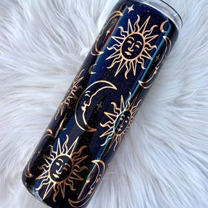 May include: A cylindrical tumbler with a dark navy blue glitter finish featuring a repeating pattern of gold-colored suns and crescent moons. The design includes detailed facial features on the suns and moons, with small stars scattered throughout.
