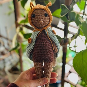 Puede incluir: Muñeca de ganchillo hecha a mano con cuerpo, piernas y cara marrones. La muñeca lleva una capucha marrón con antenas y una capa azul claro. La muñeca se sostiene en una mano sobre un fondo de plantas verdes.