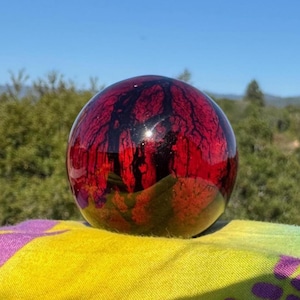 May include: A smooth, spherical object featuring a deep red and black marbled pattern throughout its surface. The sphere has a glossy, reflective finish that catches the light, creating a high-contrast appearance with dark, vein-like structures.