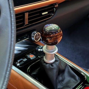 May include: Close-up of a car's interior, showcasing a wooden gear shift knob with a silver base. The knob displays a gear pattern. The interior features brown leather and black accents, including the steering wheel and dashboard.
