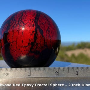 May include: A red and black epoxy fractal sphere, 2 inches in diameter, sits on a metal surface. The sphere has a glossy finish and a unique pattern. The text "Bloodwood Red Epoxy Fractal Sphere - 2 Inch Diameter" is at the bottom.