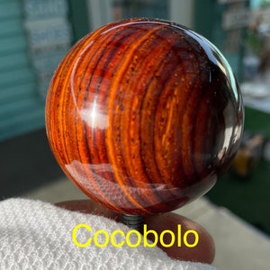May include: A round, polished cocobolo wood sphere with a glossy finish. The wood grain features a rich pattern of reddish-brown and dark brown stripes. The word "Cocobolo" is written in yellow at the bottom.