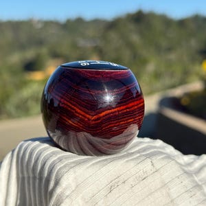 Può includere: Un pomello del cambio sferico caratterizzato da una venatura del legno lucida color rosso intenso e marrone scuro. La parte superiore del pomello presenta un inserto circolare nero piatto con lo schema delle sei marce e retromarcia inciso in bianco.