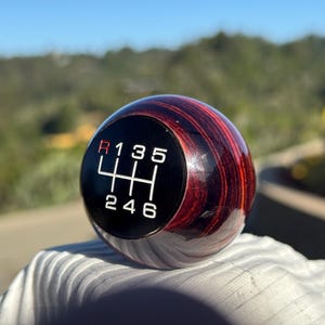 May include: A round, wooden shift knob with a dark red and brown striped pattern. The knob has a black face with white numbers and a gear shift pattern. The reverse gear is marked in red. The background is blurred.