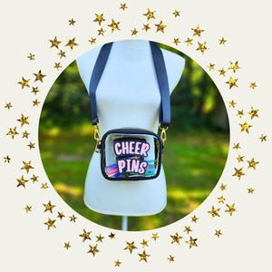 May include: A clear crossbody bag with a black zipper and strap, displaying the text "CHEER PINS" in pink and white. The bag is filled with colorful pins and accessories. The bag is displayed on a white mannequin.