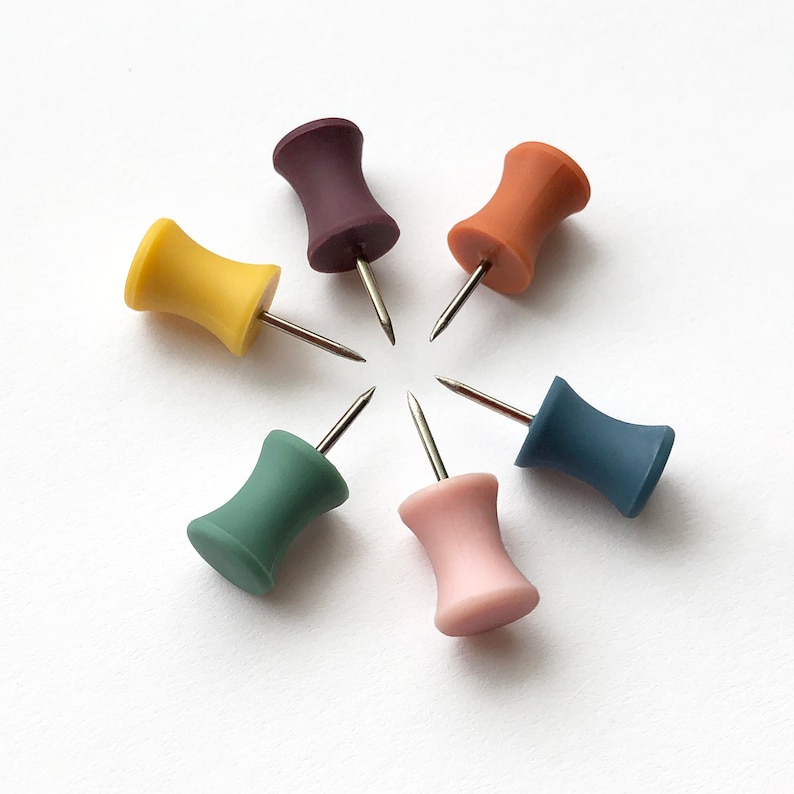 K&ouml;nnte beinhalten: Sechs bunte Stecknadeln, die in einem Kreis auf einem wei&szlig;en Hintergrund angeordnet sind. Die Nadeln sind in den Farben Gelb, Lila, Orange, Gr&uuml;n, Blau und Rosa.