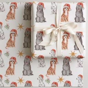 May include: A roll of white wrapping paper featuring a repeating pattern of watercolor dogs wearing red Santa hats, interspersed with green mistletoe sprigs and gold stars. A gift box wrapped in the same paper is tied with a cream satin ribbon.
