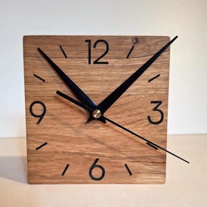 Może przedstawiać: Kwadratowy drewniany zegar z czarnymi wskazówkami i czarnymi cyframi na jasnym drewnianym tle. Widoczne są cyfry 12, 3, 6 i 9.