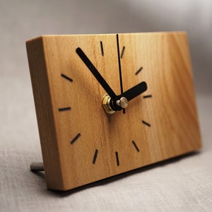 Könnte beinhalten: Eine rechteckige Holzuhr mit hellbraunem Finish. Das Zifferblatt hat schwarze Zeiger und schwarze Striche, die die Stunden markieren. Die Uhr steht auf einem kleinen Metallständer.
