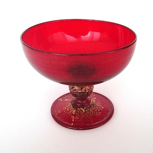 以下が含まれることがあります： 鮮やかな赤色のガラスボウル、台座付き。ボウルは丸みを帯びた形状で、光沢のある仕上がりです。ベースとステムには金色のフレークが施され、エレガントさを添えています。デザートの盛り付けや小物のディスプレイに適した装飾ボウルです。