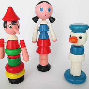 May include: Three vintage wooden toys: a Pinocchio figure with a red hat, a doll with a pink face and blue and red tiered dress, and a duck figure with a blue hat. The toys are painted in bright colors.