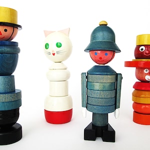 May include: Four vintage wooden stacking toys. The toys include a figure with a yellow hat, a white cat with green eyes, a figure with a blue hat, and a clown with a yellow hat. Each toy is made of painted wooden pieces.