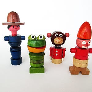 May include: Four colorful wooden toys, including a figure with a brown hat, a green frog, a monkey, and a clown. The toys are painted in bright colors and have a whimsical design, perfect for children.
