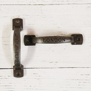 May include: Two black metal door handles with the word "LONDON" embossed on them. The handles are on a white wooden background.