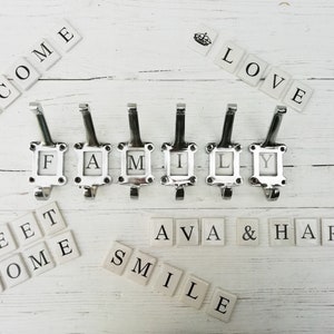 May include: White wooden wall with silver hooks spelling out the word "FAMILY".  Surrounding the hooks are white tiles spelling out the words "WELCOME", "LOVE", "SWEET HOME", "AVA & HARRY", and "SMILE".