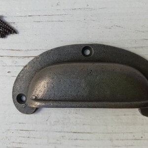 May include: A dark gray metal drawer pull with a rounded, oval shape. The pull has a smooth, slightly textured surface and two small holes for mounting screws.