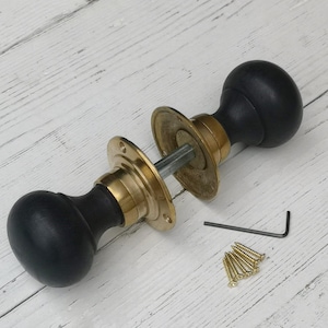 May include: A set of black wooden door knobs with brass backplates and a connecting rod. The hardware includes screws and an Allen key, presented on a white wooden surface.