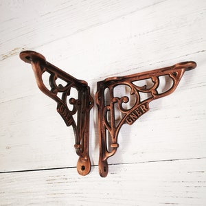 Puede incluir: Dos soportes de estante de cobre antiguo con diseños ornamentados. Cada soporte tiene la palabra "GNER" estampada.