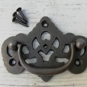 May include: A black metal drawer pull with a decorative, ornate design. The pull has a loop handle and is attached to a plate with a scalloped edge.