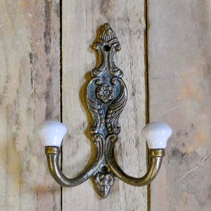 Op de afbeelding: Een dubbele haak met een decoratief, versierd ontwerp in een antieke messing afwerking. De haken hebben witte keramische eindstukken.