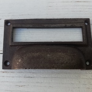 Puede incluir: Un tirador de cajón rústico de hierro fundido con un recorte rectangular y una manija redondeada y empotrada. El tirador es de color marrón oscuro con un acabado desgastado.