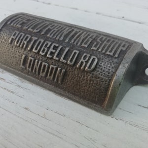 May include: A weathered metal bottle opener, embossed with "THE OLD PAINTING SHOP, PORTOBELLO RD, LONDON". The opener has a textured finish and a hole for hanging, a classic design.