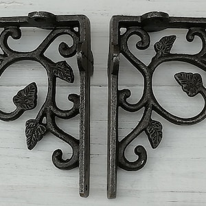Könnte beinhalten: Zwei schwarze Metall-Regalhalterungen mit einem dekorativen floralen Design. Die Halterungen sind aus Gusseisen und haben eine rustikale Oberfläche.
