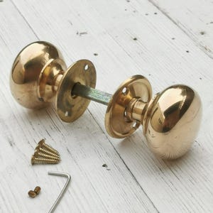 Op de afbeelding: Een paar gepolijste messing deurknoppen met bijpassende achterplaten en een verbindingsstang. De set bevat schroeven en een inbussleutel, allemaal weergegeven op een lichtgekleurd houten oppervlak. De knoppen hebben een klassiek, afgerond ontwerp.