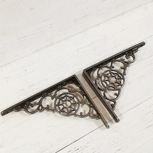 Op de afbeelding: Twee sierlijke metalen plankdragers met een decoratief, ingewikkeld ontwerp. De dragers zijn donkerbruin van kleur en hebben een verweerde afwerking.