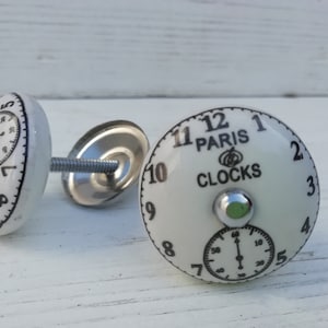 Peut inclure: Deux boutons de cabinet en céramique blanche avec un design de cadran d'horloge. Un bouton porte le texte "PARIS CLOCKS" et l'autre bouton a un cadran d'horloge plus petit.