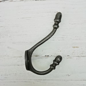 Pode incluir: Um gancho de metal prateado com um design de bolota na parte superior e inferior. O gancho está montado em uma superfície de madeira branca.