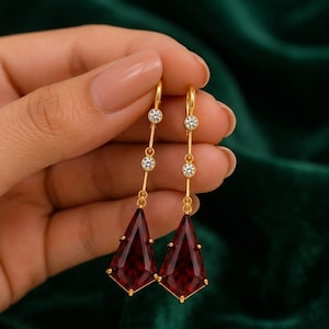 May include: A pair of gold drop earrings featuring two small round clear stones set along a thin vertical chain, terminating in a large kite-shaped deep red gemstone. The earrings are constructed with a polished gold metal finish and secure hook backings.