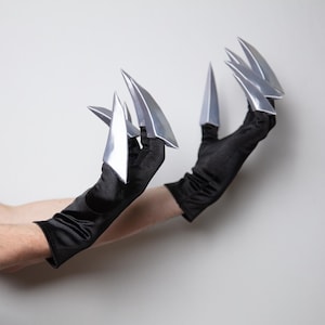 May include: Two hands wearing black gloves with silver, claw-shaped attachments. The claws are sharp and metallic, reflecting light. The gloves are long, made of a dark, smooth material. The background is a plain white, highlighting the accessories.