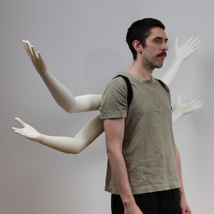 Puede incluir: Una persona con una camisa verde y un arnés negro, frente a brazos de maniquí blancos. Los brazos del maniquí están colocados para que parezca que salen de la espalda de la persona.