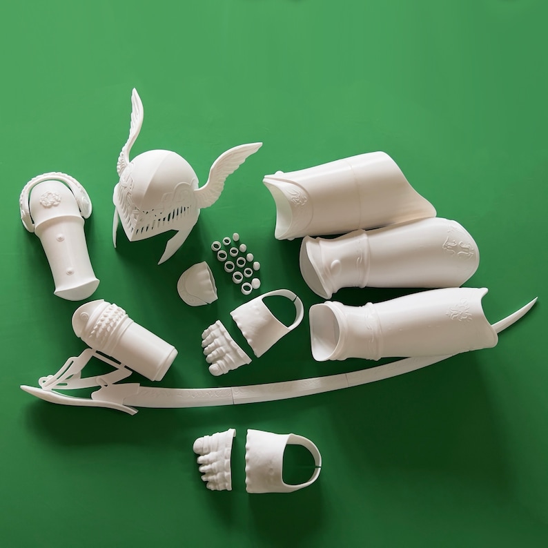 May include: White plastic 3D printed model kit of a knight's armor. The kit includes a helmet, gauntlets, leg armor, and a sword. The pieces are laid out on a green surface.