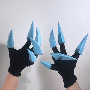 Pode incluir: Um par de luvas pretas com extensões em forma de garra azul claro. As pontas das garras são pontiagudas e estendem-se das pontas dos dedos, criando um efeito visual marcante. As luvas parecem ser feitas de um material macio e flexível.