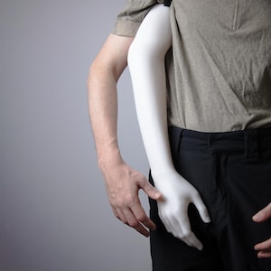 May include: A white prosthetic arm is held by a person wearing a gray shirt and black pants. The prosthetic arm is smooth and realistic, with a hand in a relaxed position. The background is a solid gray.
