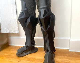 May include: A pair of black knee-high leg armor with a geometric design. The armor is made of a hard, durable material and has a sleek, futuristic look.