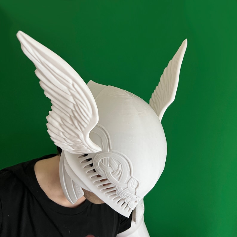 May include: White plastic helmet with wings, resembling a winged helmet from a fantasy movie or video game. The helmet has intricate details and a textured surface.