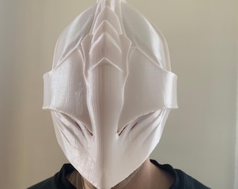 Könnte beinhalten: Eine glatte, cremeweiße Vollmaske mit einem markanten Mittelsteg und kantigen, flügelartigen Seitenerweiterungen. Die Maske hat ein geschichtetes, segmentiertes Aussehen mit einer spitzen Oberseite und einer glatten, geformten Oberflächenstruktur.