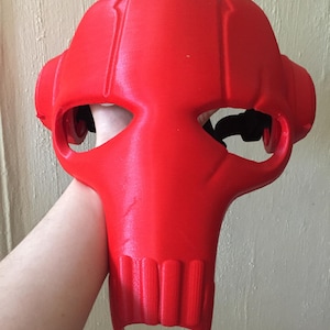 May include: A red plastic mask with a skull-like design. The mask has two eye holes and a mouth opening. The mask is 3D printed and has a textured surface.