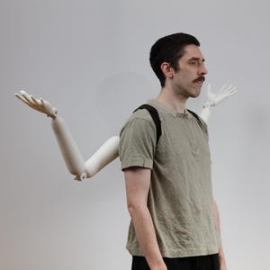 Może przedstawiać: Osoba w jasnozielonej koszuli i czarnych spodniach z białym, przegubowym ramieniem przymocowanym do pleców. Ramię jest ustawione z otwartą dłonią, jakby gestykulowało.