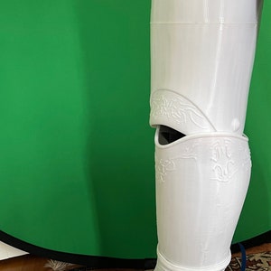 May include: White 3D printed leg armor with intricate details and a decorative design. The armor covers the lower leg and foot.