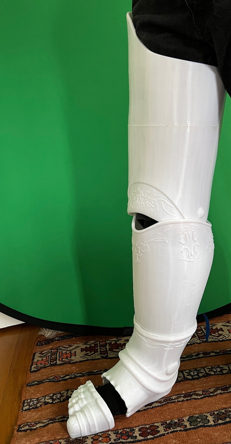 May include: White 3D printed leg armor with intricate details and a decorative design. The armor covers the lower leg and foot.