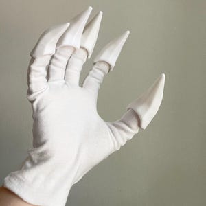 May include: A white fabric glove featuring five rigid, pointed white claw attachments secured to the fingertips. The glove covers the hand and wrist, with each finger and the thumb capped by a smooth, tapered, claw-like extension in matching white.