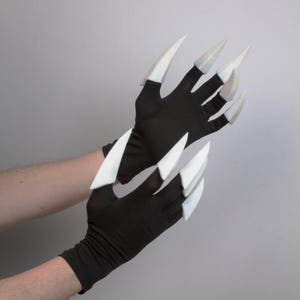 Pode incluir: Luvas pretas com grandes extensões brancas em forma de garra. As luvas são feitas de um material elástico e as garras são pontiagudas. As luvas são projetadas para se assemelhar a garras de animais.